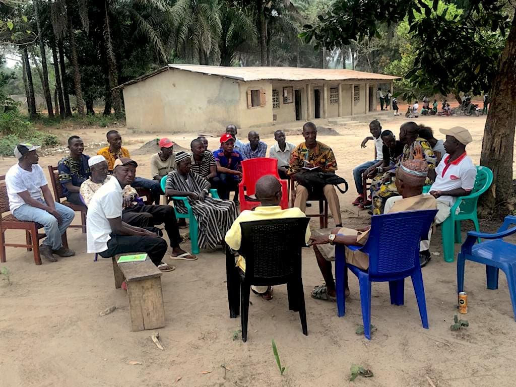 Stakeholdermeeting für die neue Schule in Barbara, Sierra Leone. Eine große Gruppe Menschen sitzt auf Plastikstühlen im Kreis vor dem alten Schulgebäude in Barbara und bespricht das neue Bauvorhaben.